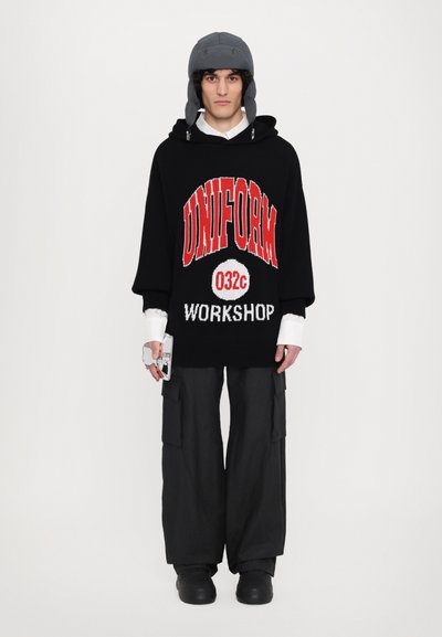 Schwarzer oversized Hoodie mit dem Schriftzug "UNIFORM 032c WORKSHOP" in rot und weiß, kombiniert mit weiten schwarzen Cargo-Hosen und einer grauen Mütze.