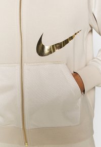 Beige zip-up hoodie med en stor gul Nike-logotyp på bröstet, med texturerat tyg och en ficka fram på vardera sidan.