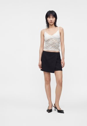 Femme debout de face, portant un caraco en dentelle blanche, une mini-jupe noire asymétrique et des talons noirs sur un fond uni et clair.