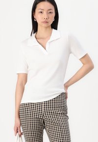 Hvid kortærmet polo-shirt med krave, glat tekstur, parret med sorte og beige ternede bukser. Rent, enkelt design.