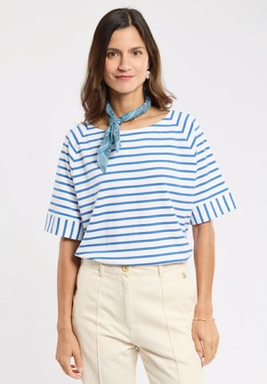 Femme aux cheveux bruns mi-longs portant une chemise blanche à rayures bleues, un pantalon beige taille haute et un foulard bleu autour du cou.