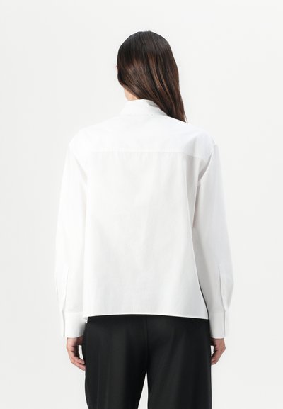 White button-up shirt featuring long sleeves, a straight cut, and back seam detailing, paired with black trousers. Smooth fabric texture.