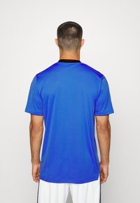 Krátký rukáv modrého sportovního dresu s černým límečkem. Hladká textura, uvolněný střih a viditelné švy podél ramenních švů.