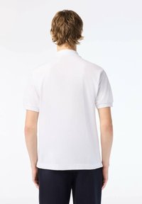 Young person with short brown hair wearing a plain white short-sleeve polo shirt and dark pants, standing facing away on a white background.