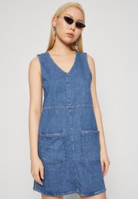 Denim haljina s V-izrezom, bez rukava, koja ima dva velika prednja džepa i kontrastne šavove. Boja: srednje plava denim tekstura.