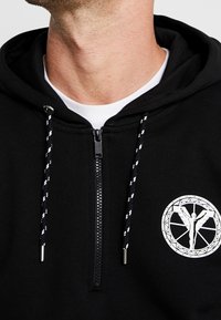 Felpa nera con zip, coulisse bianche e un logo circolare stampato che presenta una figura all'interno di design geometrici. Materiale in cotone.