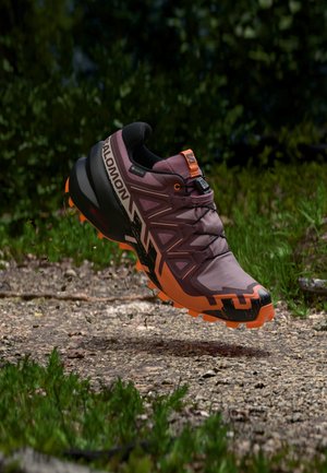 Παπούτσι για trail running με πορτοκαλί και μωβ σχεδιασμό, παχιά σόλα με πέλμα και σήμανση Salomon, να αιωρείται πάνω από μονοπάτι με χαλίκι και γρασίδι.