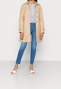 Beige trenčkot s límcem, s knoflíkovou přední částí a ohrnutými rukávy, nošený přes modro-bílý vzorovaný top a modré džíny. Bílé sandály na podpatku.