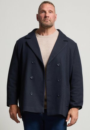 Homme aux cheveux courts et à la barbe, portant un pull beige sous un manteau croisé bleu foncé, debout devant un fond uni.