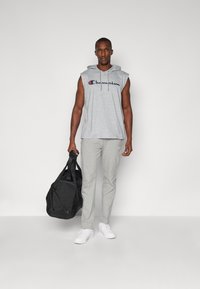 Grijze mouwloze hoodie met 'Champion' logo, grijze taps toelopende broek, witte sneakers en een zwarte sporttas, tegen een effen achtergrond.