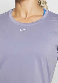 Tričko s krátkým rukávem, kulatým výstřihem a sportovním střihem v světle levandulové barvě s hladkou texturou a jemným leskem; na hrudi se nachází malé bílé logo Nike.