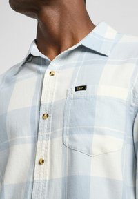 Camisa de cuadros azul claro y blanca con cuello abotonado, botones dorados y un bolsillo en el pecho que presenta una etiqueta negra. Textura de tela suave.