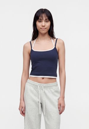 Junge Frau mit langen dunklen Haaren, die ein marineblaues Crop-Top mit weißem Rand und hellgraue Jogginghose vor einem einfarbigen weißen Hintergrund trägt.