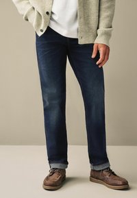 Donkerdenim jeans met een klassieke pasvorm, gedragen met een lichtgrijze cardi en bruine leren schoenen, met opgerolde mouwen en minimale slijtage.