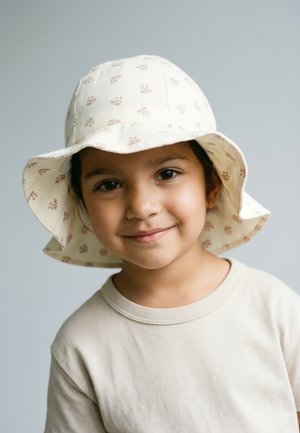 Junges Kind lächelt, trägt einen hellbeigen floralen Fischerhut und ein passendes beiges Hemd, vor einem schlichten grauen Hintergrund.