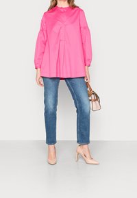 Blusa lunga a maniche con collo alto di un vivace colore rosa, abbinata a jeans blu e tacchi nudi, mentre tiene una piccola borsa a tracolla.