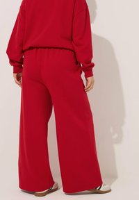 Rode wijdvallende sweatpants met elastische tailleband, zachte stof en contrasterende schoenen zichtbaar aan de onderkant. Geen opvallende patronen of accenten.