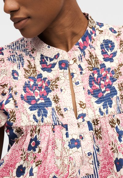 Gros plan d'une personne portant un chemisier à motifs floraux rose, bleu et brun, avec une encolure en trou de serrure boutonnée et un détail froncé sur la poitrine.