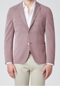 Blazer rosso con texture, dal taglio sartoriale, dotato di due tasche frontali e chiusura con un solo bottone, abbinato a una camicia bianca sotto.