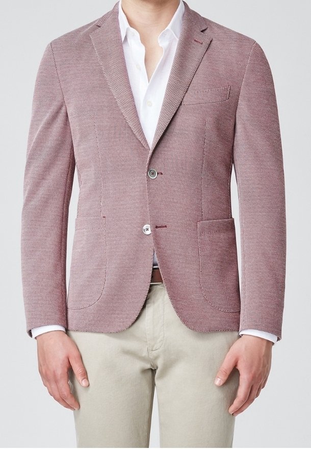 Blazer rosso con texture, dal taglio sartoriale, dotato di due tasche frontali e chiusura con un solo bottone, abbinato a una camicia bianca sotto.