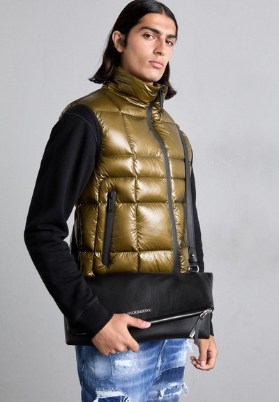 Gilet vert olive rembourré avec un col haut sur une chemise à manches longues noire. Sac en cuir noir avec fermeture éclair tenu dans une main. Jeans usés.