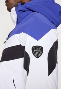 Syntetická bunda s farebnými blokmi v modrej, bielej a čiernej farbe. Má predný zips a vyšívané logo EA7 na rukáve.