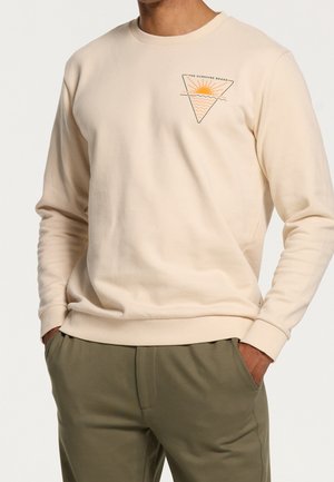 Beige sweatshirt met lange mouwen met een klein driehoekig logo met zon en golven en de tekst "The Sunshine Brand" op de borst, gedragen met olijfgroene broek.