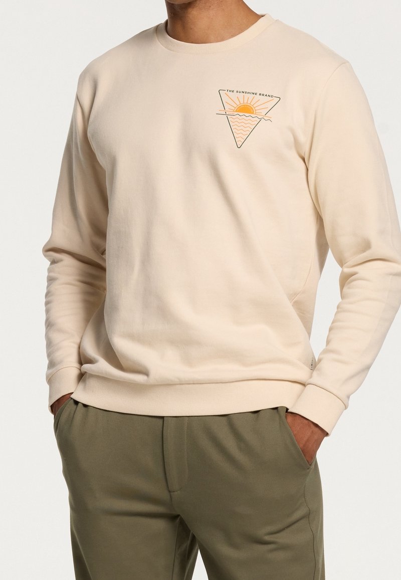 Sweat-shirt beige à manches longues avec un petit logo triangulaire représentant un soleil et des vagues, et le texte "The Sunshine Brand" sur la poitrine, porté avec un pantalon olive.