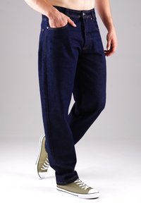 Dunkelblaue Denim-Jeans mit lockerer Passform, fünf Taschen und kontrastierenden Nähten, kombiniert mit hellgrünen Sneakers und weißen Gummiakzenten.