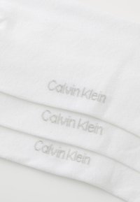 Hvite bomullssokker med en myk tekstur, med en subtil grå "Calvin Klein"-logo brodert nær toppen. Tre sokker synlige.