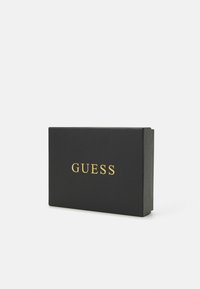 Boîte rectangulaire noire avec le logo doré "GUESS" à l'avant. Texture lisse, léger éclat, design refermable, taille compacte.