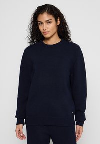 Marineblauer gestrickter Pullover mit gerippten Bündchen und Saum, der einen Rundhalsausschnitt und ein kleines metallisches Logo-Detail am unteren Ende aufweist.