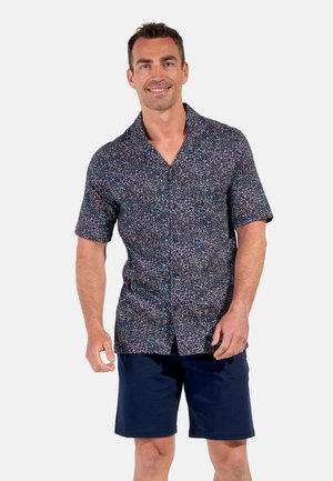 Mand smilende iført en kortærmet blomsterprint skjorte med knapper og marineblå shorts, stående mod en simpel lys baggrund.