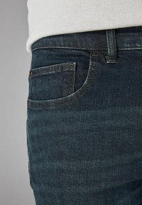 Primer plano de un bolsillo de jeans de mezclilla azul oscuro con costuras visibles y una prenda blanca metida en la cinturilla.
