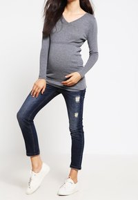 Graues langärmliges V-Ausschnitt-Maternity-Top mit figurbetontem Schnitt, kombiniert mit dunkelblauen Jeans aus Denim mit Destroyed-Details und weißen Sneakers.