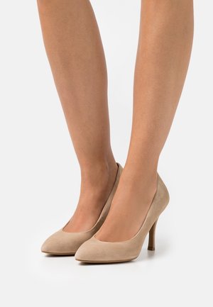 Femme portant des escarpins beiges en daim à bout pointu, se tenant sur une surface blanche.