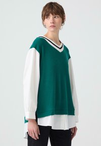 Gilè a coste di colore verde con scollo a V e colletto a righe, indossato sopra una camicia bianca. Vestibilità ampia, presenta maniche lunghe e una silhouette rilassata.