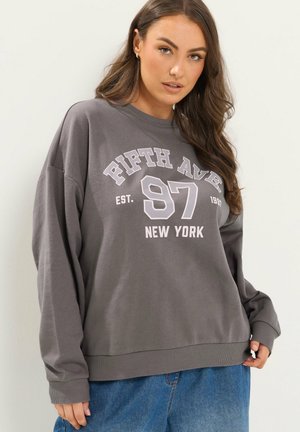 Kvinde med langt brunt hår har en løs grå sweatshirt med "Fifth Ave 97 New York" i hvid tekst og blå jeans.