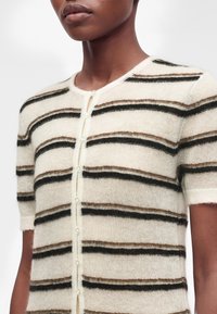 Personne portant un cardigan à manches courtes boutonné avec des rayures horizontales noires et marron sur un fond écru.