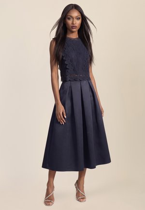 Frau mit langen glatten Haaren, die ein ärmelloses marineblaues Spitzenoberteil und einen plissierten Midirock trägt, kombiniert mit silbernen Sandalen mit hohen Absätzen.