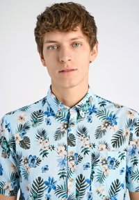 Korte mouwen, knopsluiting shirt van lichtblauwe stof met tropische bloem- en bladenpatronen in tinten groen, blauw en beige.