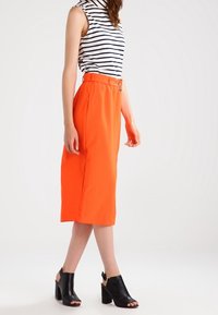 Orange Midi-Rock mit tailliertem Gürtel, kombiniert mit einem gestreiften ärmellosen Oberteil und schwarzen Blockabsatzsandalen. Glatte Textur und passgenauer Schnitt.