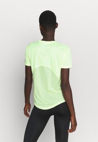 Osoba z krótkimi włosami nosząca jasnorudą koszulkę sportową z krótkim rękawem i siatkowym tyłem oraz czarne legginsy, stojąca plecami do obiektywu na jednolitym tle.