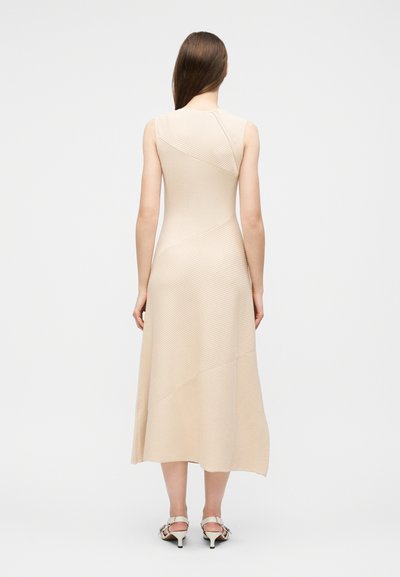 Femme aux longs cheveux bruns portant une robe midi beige sans manches et des sandales blanches à talons, debout de dos sur un fond blanc uni.