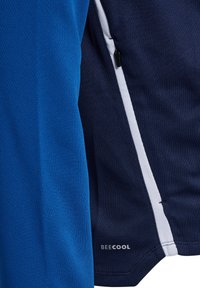 Close-up van blauw en donker marine sportkledingstof met een witte streep en een ritssluiting nabij de naad, met de tekst "BEECOOL" aan de onderzoom.