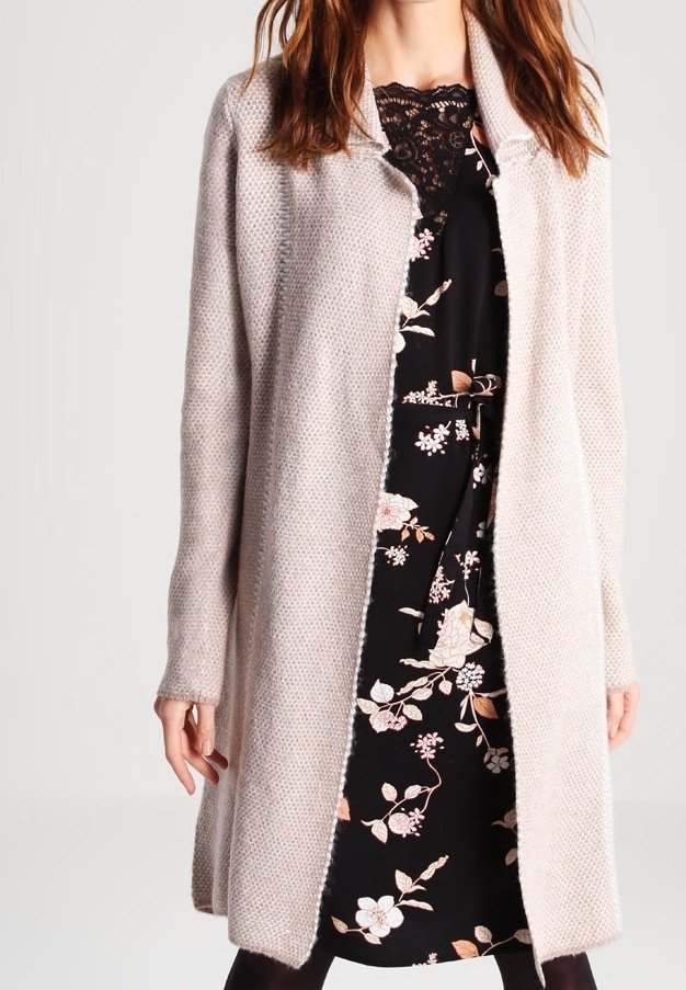 Cardigan beige clair, texture tricotée douce, devant ouvert, manches longues, portant une robe noire à motifs floraux en contraste en dessous.