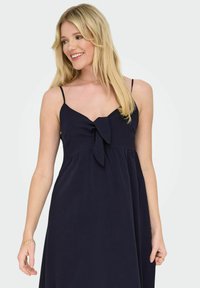 Robe bleu marine à fines bretelles, avec un décolleté en V et un détail de nœud au niveau de la poitrine. Tissu léger et fluide avec une coupe décontractée.