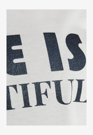 Primo piano di un tessuto bianco con testo nero glitterato che mostra parole parziali "E IS" e "TIFUL" in carattere grassetto.