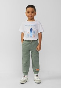Grüne Sweatpants mit strukturierter Oberfläche, kombiniert mit einem weißen T-Shirt, das Skateboardgrafiken in Blau und Orange zeigt. Weiße Sneakers.