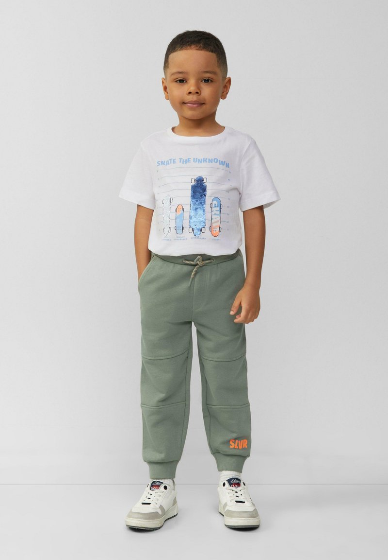 Grüne Sweatpants mit strukturierter Oberfläche, kombiniert mit einem weißen T-Shirt, das Skateboardgrafiken in Blau und Orange zeigt. Weiße Sneakers.